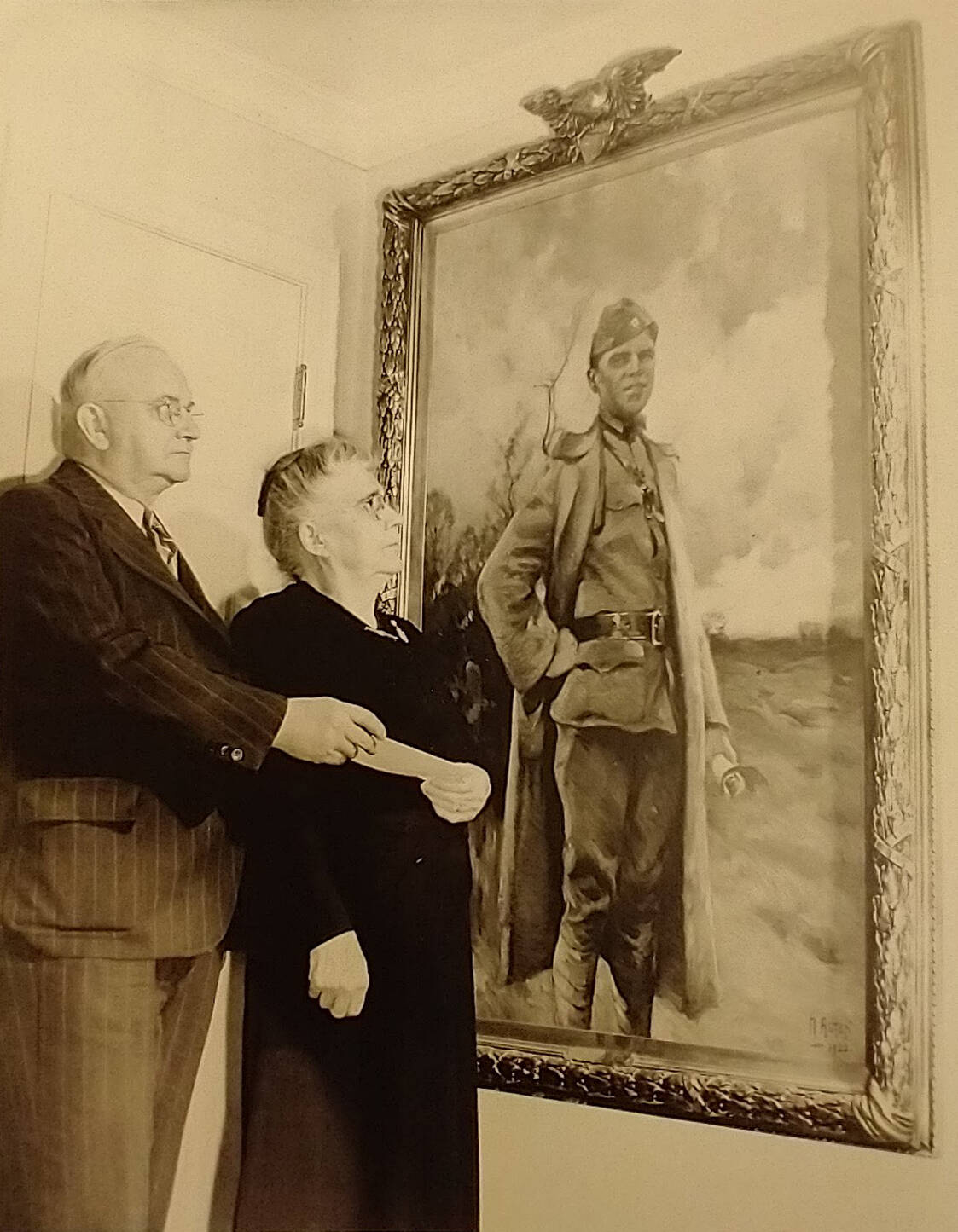 Photograph of a man and woman standing in front of a painting hanging on a wall of a soldier in uniform.